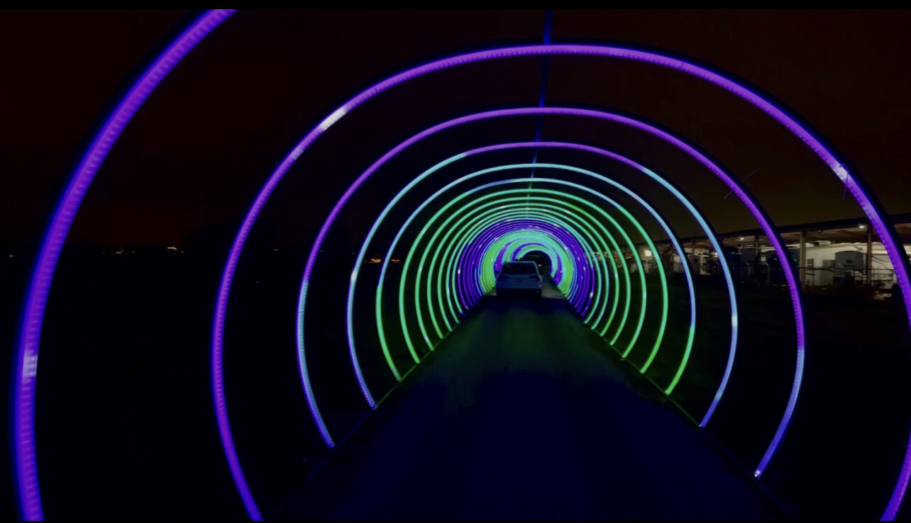 A car driving beneath a glowing tunnel of colorful holiday lights